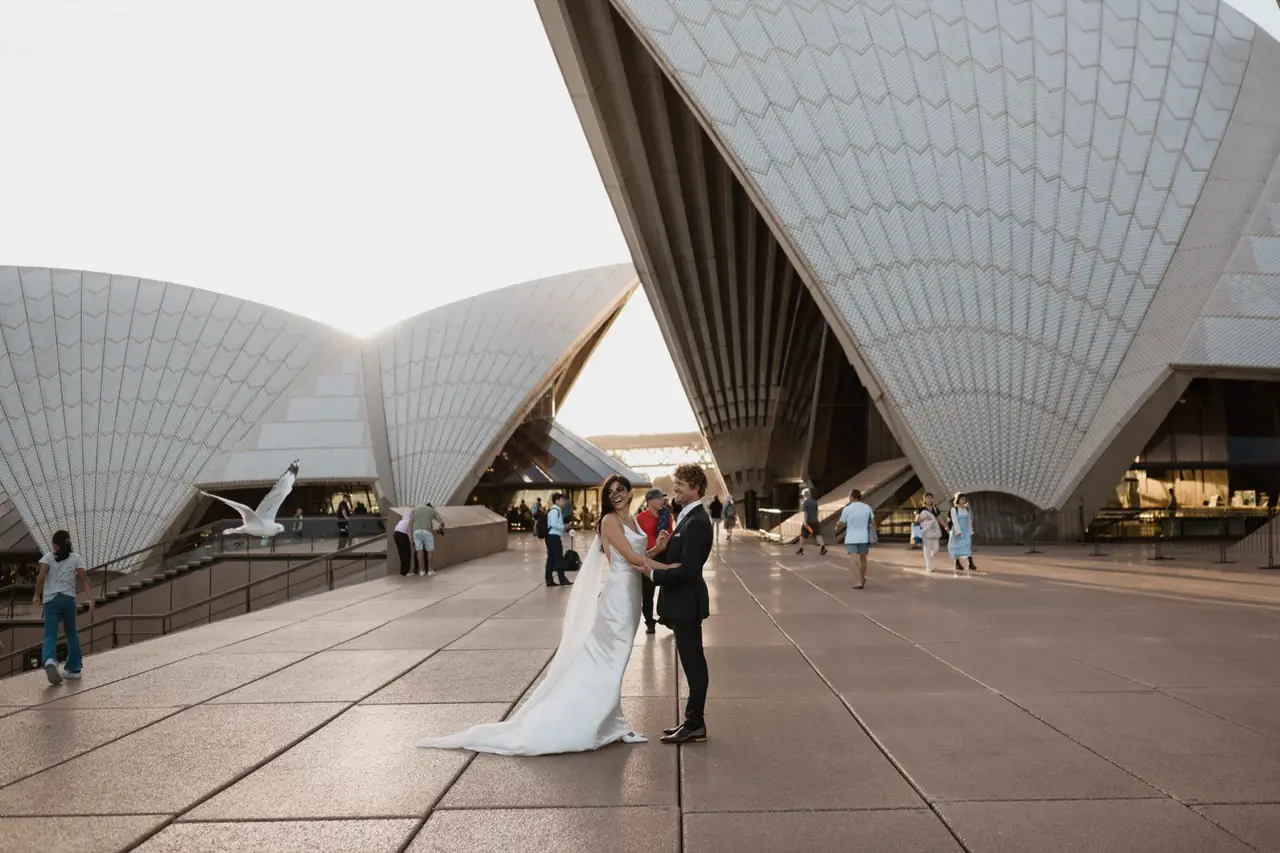 A newly married couple standing beneath the sails of the Sydney Opera House.